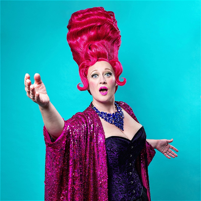 female comedian with pink beehive wig, purple corset, and fuchsia sequin kaftan is reaching toward the camera