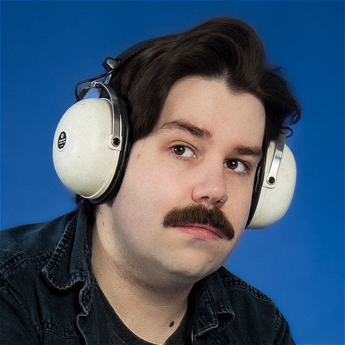 Male comedian with brown hair and a moustache wears vintage headphones on a blue background
