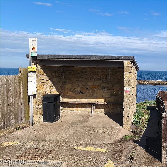 A simple, open-sided stone bus stop shelter located directly beside the sea under a bright blue sky. A black bin stands next to the wooden bench inside the shelter