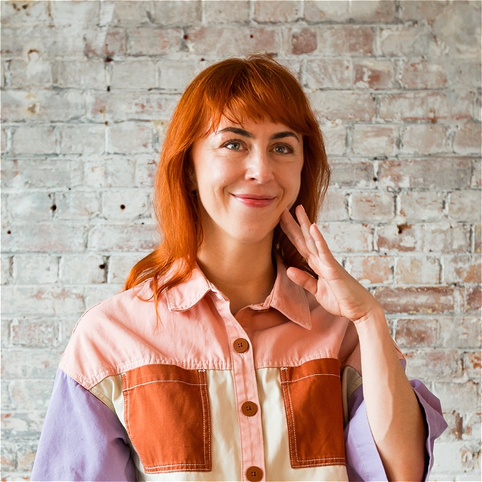 female comedian with red hair standing in front of brick wall