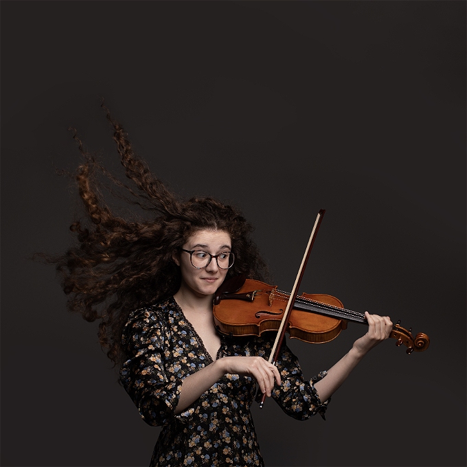 Female comedian with brown hair playing the violin in front of a grey background.