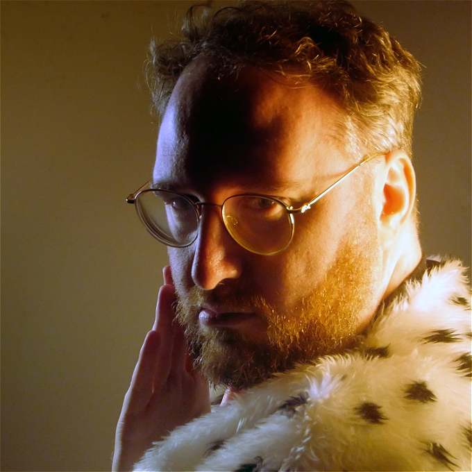 Male comedian in faux fur robes turns coquettishly towards the camera with dead eyes
