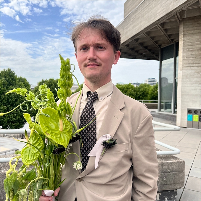 White male comedian in tan suit holding weird flowers
