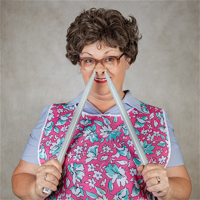 Female Comedian, Curly Brown Hair, Glasses and wearing a Tabard with Two Giant Knitting Needles touching her Nostrils