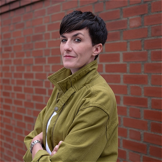 Female comedian wearing a green jacket, standing in front of a brick wall