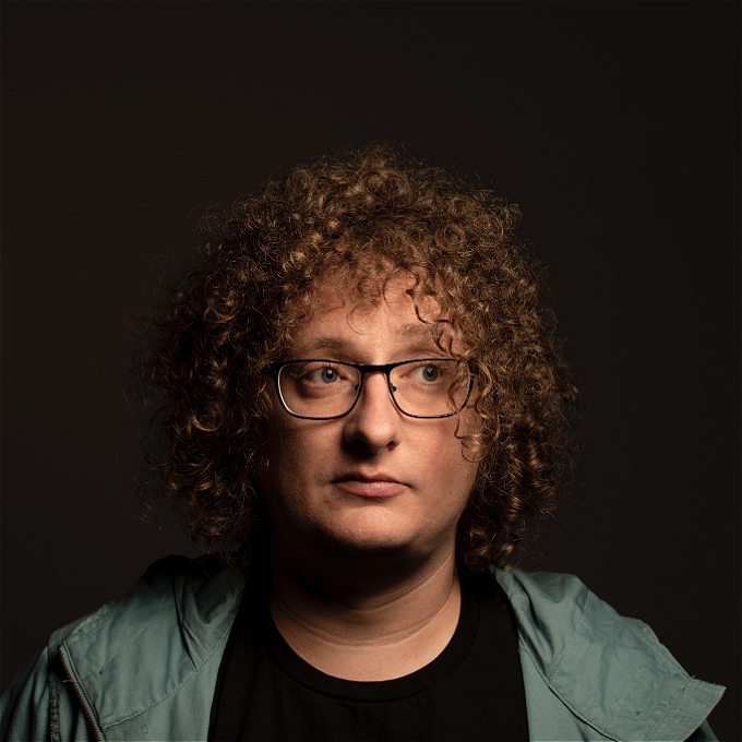 Head shot cut off at shoulders of male comedian with curly long hair and glasses, wearing a black top with a light blue jacket
