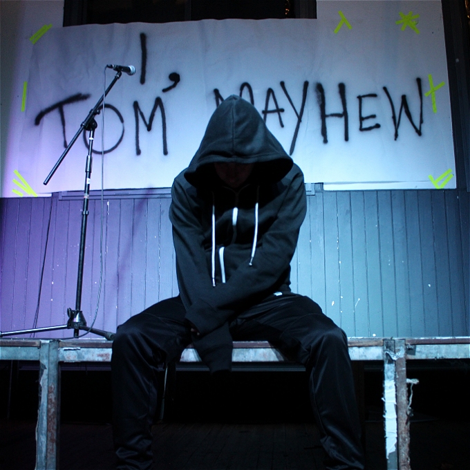 Male comedian wearing a black hoody with the hood up, sitting on a stage looking down