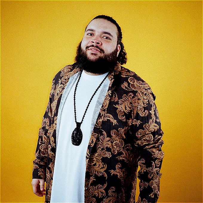 A man with a full beard and tied-back curly hair is standing against a bright yellow background. He is wearing a white t-shirt, a long patterned jacket with ornate gold and brown designs, and a long black beaded necklace with a large pendant. He is looking slightly upward with a relaxed expression