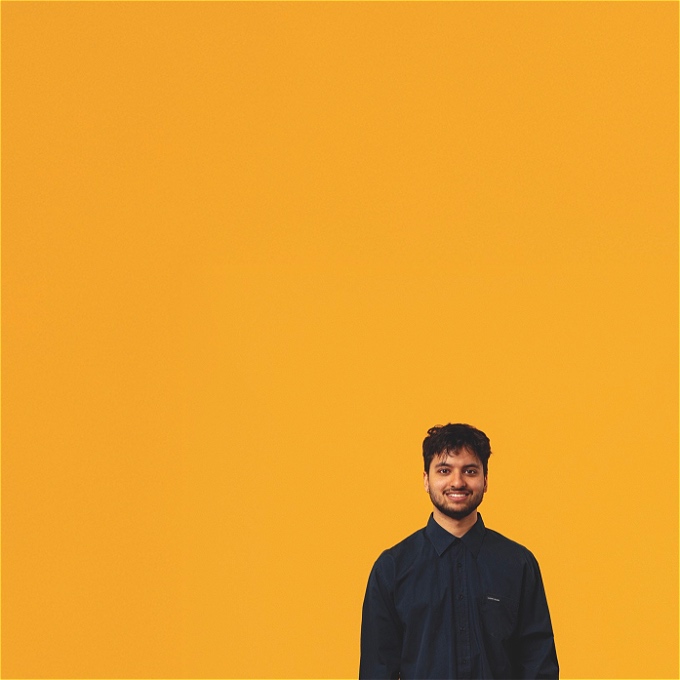 The top half of a male British Indian comedian sticks out the bottom of the picture, he looks small against a large, blank orange background
