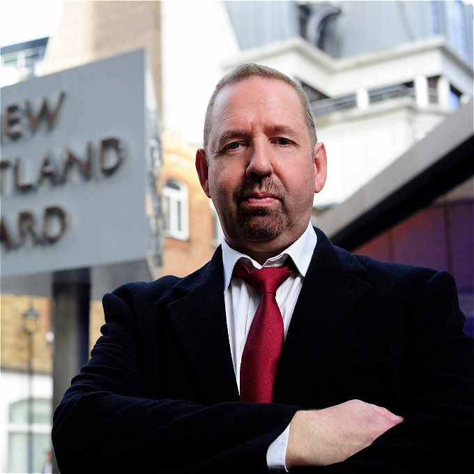 Comedian Alfie Moore, wearing a black suit, white shirt and red tie, standing with arms folded in front of the New Scotland Yard sign