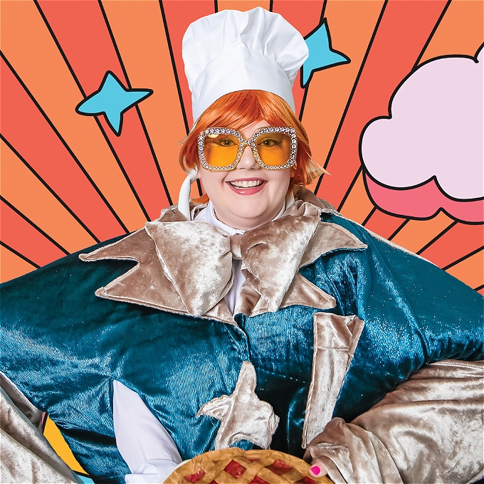 Female comedian in lovely orange short haired wig and giant chefs hat also dons yellow bejewelled glasses, a giant turquoise and gold suit jacket which spans a 40 inch shoulder, and is holding a freshly handmade apple and raspberry pie.