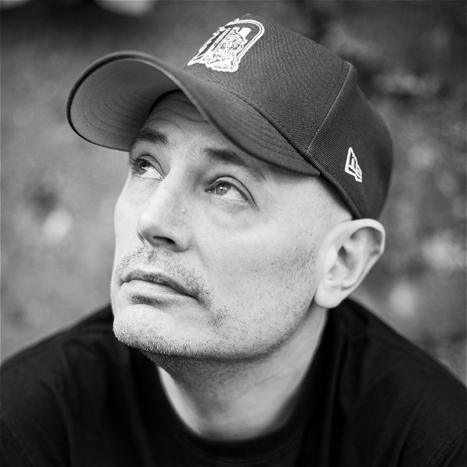 Male comedian in black and white looks away from the camera while wearing a baseball cap.