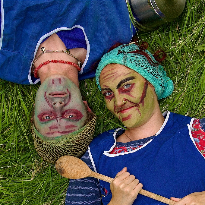 Two gruesome dinnerladies lie on the grass, looking upwards into the camera. One dinnerlady has a green face, holds a wooden spoon and is smiling. The other dinnerlady is upside down with a blue face and is frowning.