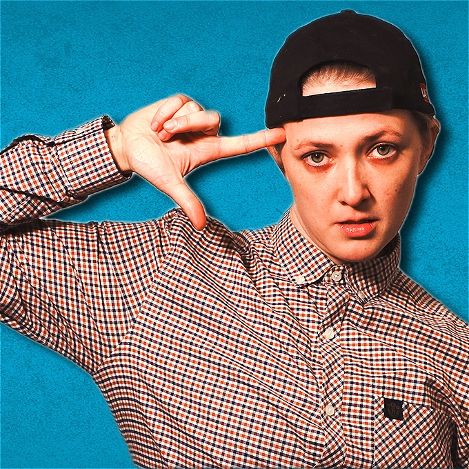 Female comedian in a checkered shirt and backwards black cap, dressed as a man, pointing her index finger to her head.