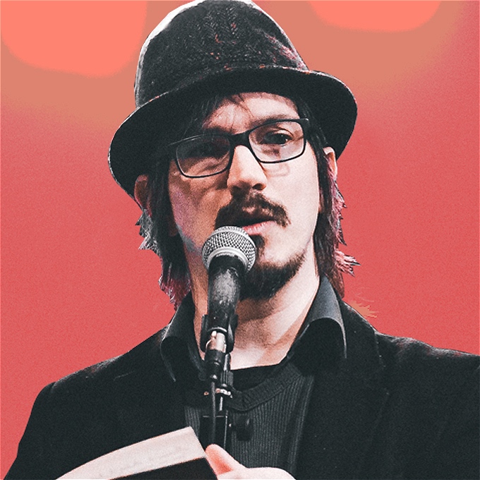 A devastatingly handsome chap in a tweed hat stands at a microphone and holds a book