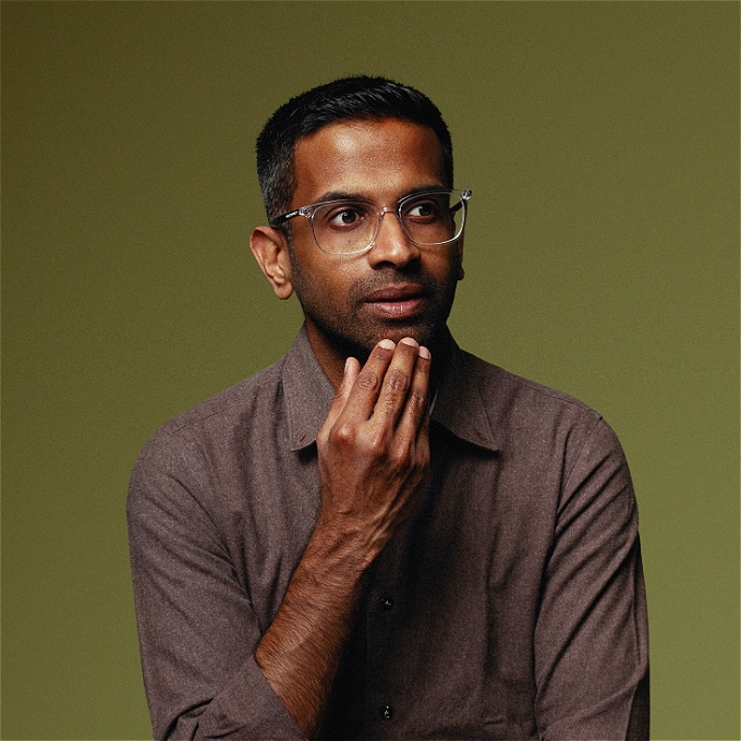 Comedian with black hair stroking chin looking pensive in green background
