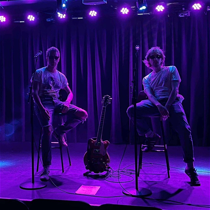 Two men with sunglasses sitting on stools either side of a guitar in purple stage light