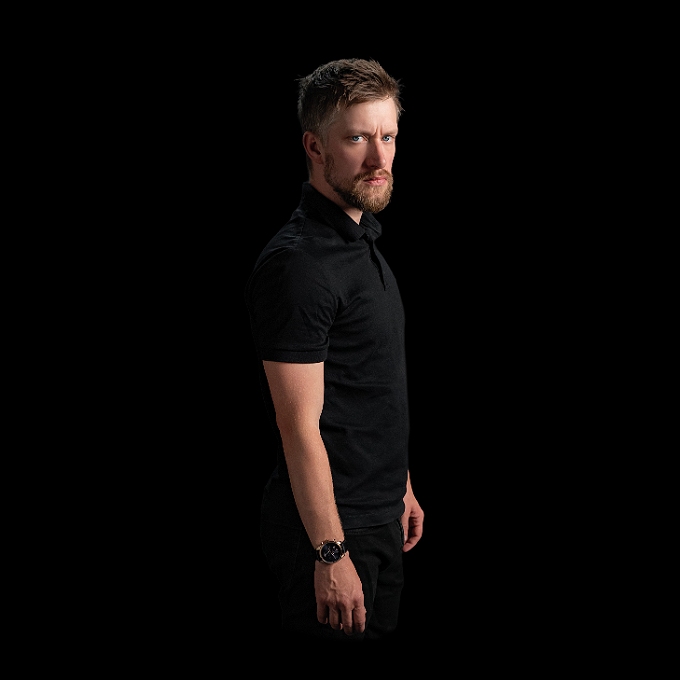 Male comedian looking to the camera, set against a black background.