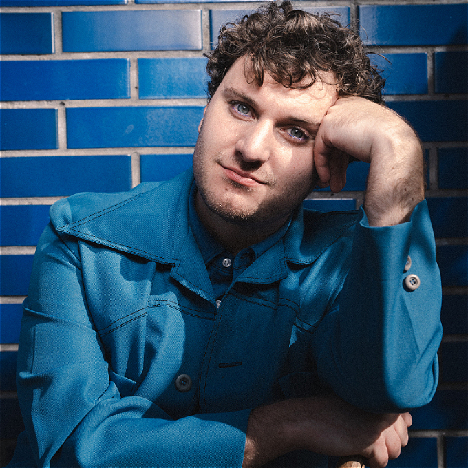 Jonathan leans on his arm whilst wearing a blue suit, blue shirt against a tiled blue background