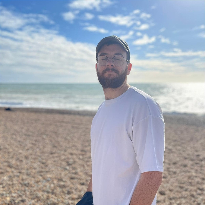 Male comedian Rich Spalding stands side on on a beach in Brighton. He's looking at the camera but has blinked so his eyes are closed. The sea is behind him.