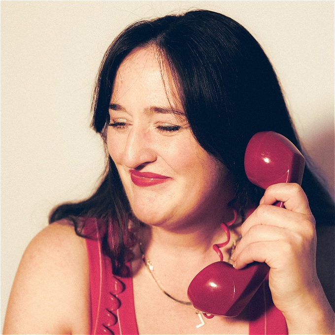 Female comedian with dark hair laughs with her eyes squinting while holding a red retro rotary telephone to her ear