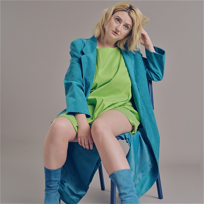 Female comedian with dyed blonde hair sits in a chair looking SUPER hot