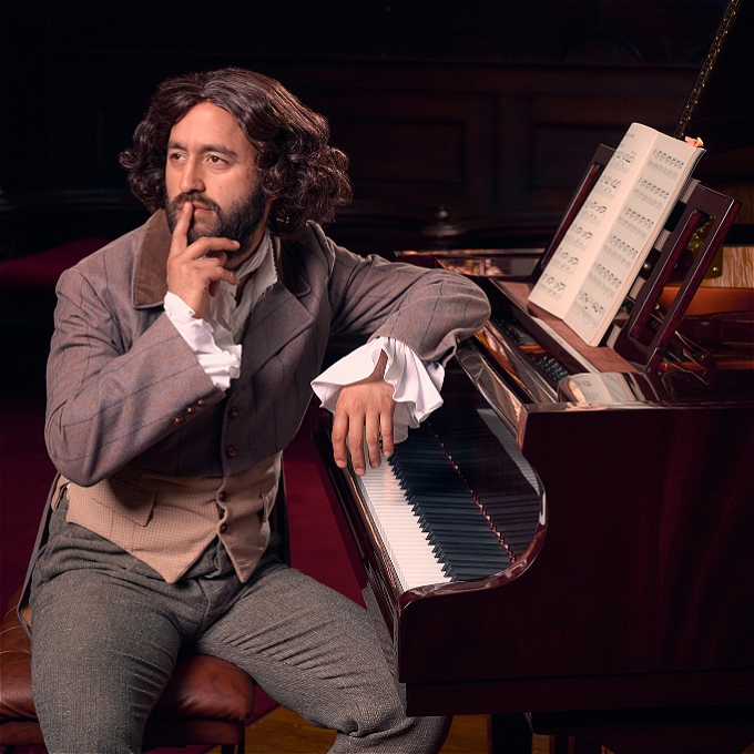 A man sitting at a grand piano, wearing a coat tail jacket and a 19th century wig