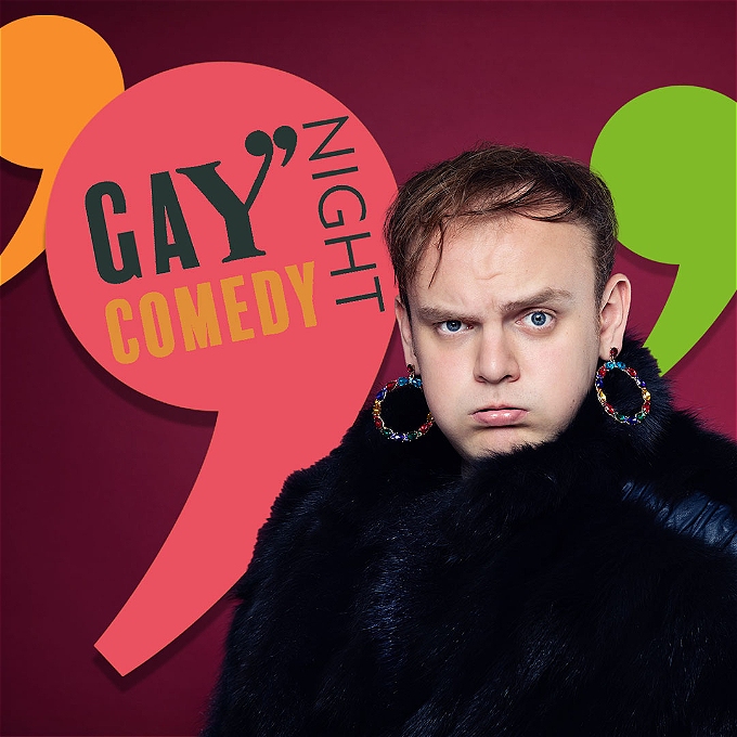 Comedian Andrew White wears a fluffy black coat and colourful hoop earrings in front of the multi-coloured Gay Comedy Night logos