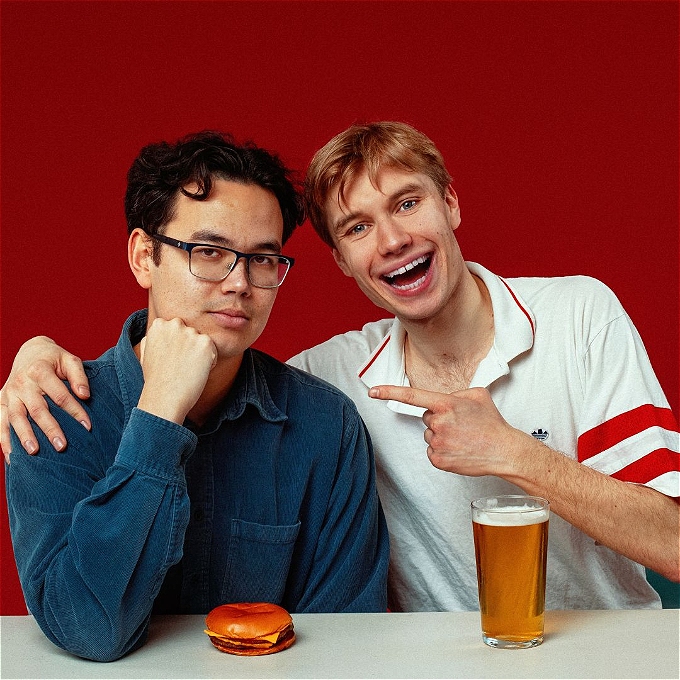 Two comedians, one in a blue shirt and one in a white sit above a burger and a pint