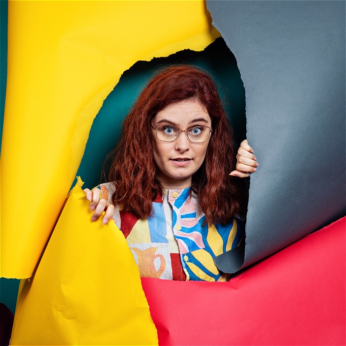 Adele is emerging from colourful torn paper (navy, red and yellow.) she has dark red hair and glasses and is wearing a bright top.