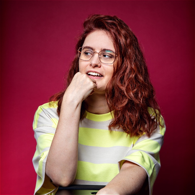 Female comedian with red/brown hair and glasses looks off camera in front of burgundy background