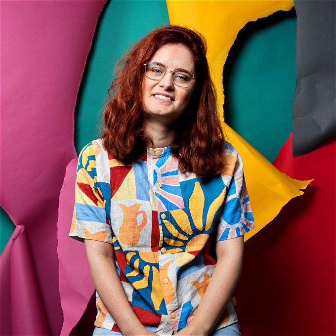 Female comedian with dark red hair and glasses smiling in front of a colourful background