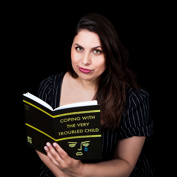 Brown female comedian looks to the camera, holding a book with the title ‘Coping With The Very Troubled Child’.