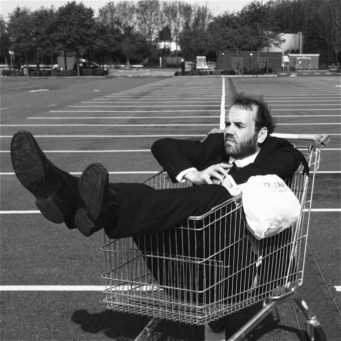 black and white photo of a male comedian in his early 40s sat in a shopping trolley looking sad holding a dicta phone. Flopped over the side of the trolley is a Somerfield carrier bag.