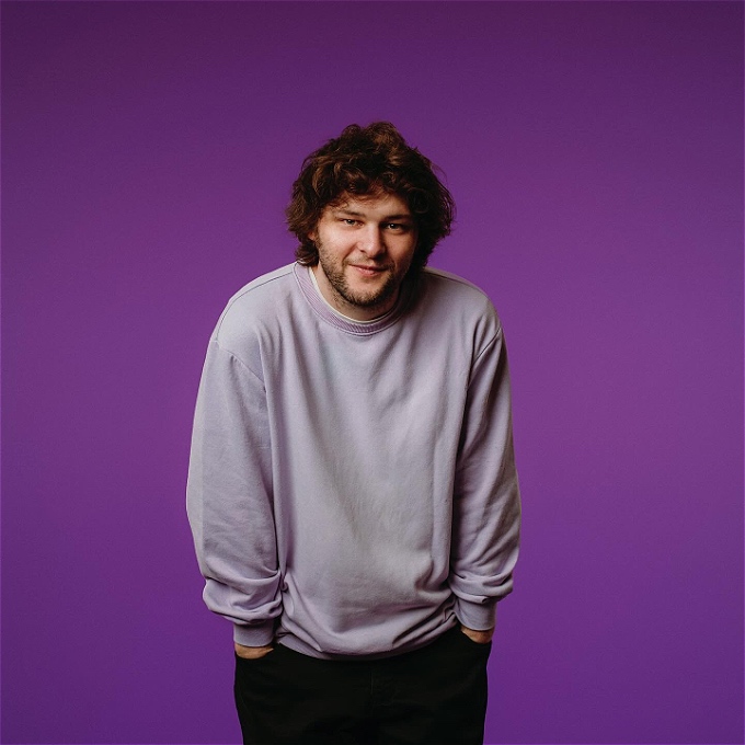 A photo of comedian Jake Baker in his favourite lilac sweater (he no longer wears it as often as he once did having, since this photo was taken, spilled hoisin sauce on it that has left a very noticeable stain) on a purple background.