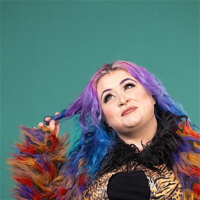 A picture of a female comedian with blue/purple hair and a bright furry coat on and leopard top looking wistfully into the distance in front of a turquoise background.