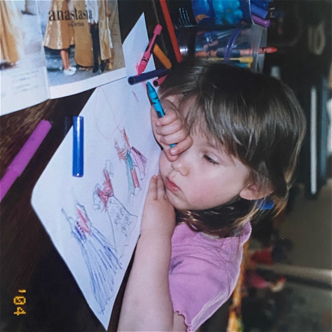 Young girl with brown hair slumps sleepily over a colouring book while holding a crayon in one hand.