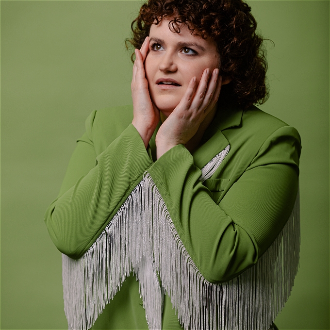 Comedian with brown curly hair in a green blazer with white fringe looking up, concerned, with her hands around her face