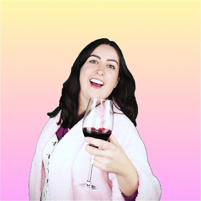 Female comedian with long brown hair in a pink dressing gown smiling while holding a glass of red wine in front of a gradient yellow to pink background
