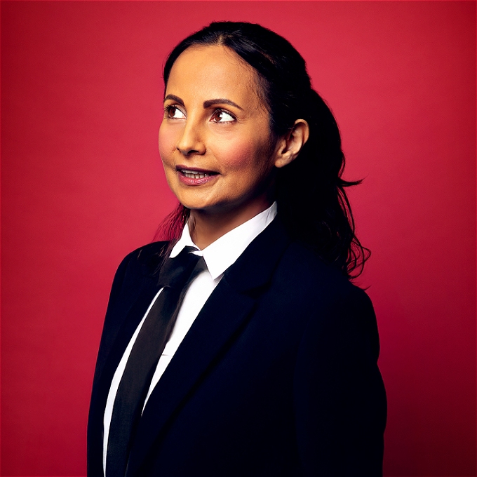 Female comedian in a suit and tie looking to the top left on a red background
