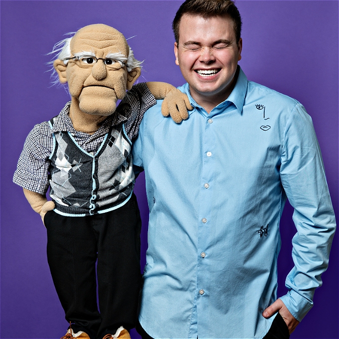 Male ventriloquist in blue shirt, laughing and holding a puppet grandad figure.