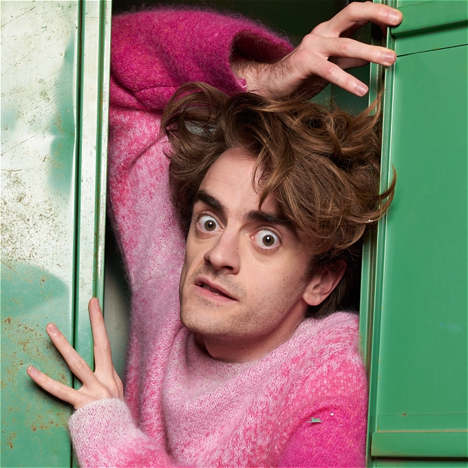 A comedian with wild hair bursts out of a green locker wearing a fluffy pink jumper