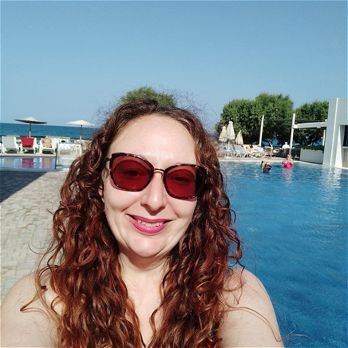 Female comedian smiling wearing sunglasses with pool and beach in background with blue sky n sunshine