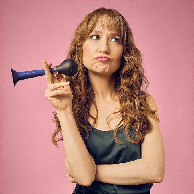 Female comedian with long ginger hair pouts and holds a blue plastic horn, standing against a pink background