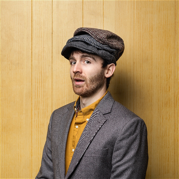 Image of headliner of the comedy show, comedian Ian Smith, wearing a grey jacket, ochre shirt, and three flat caps.