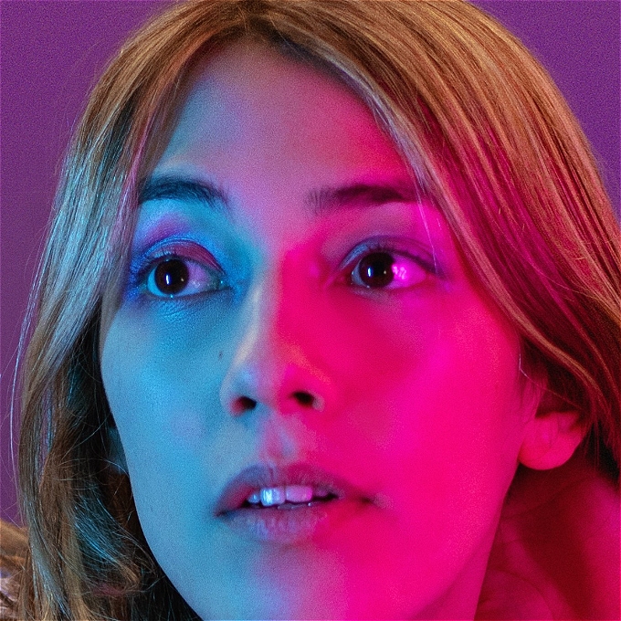 Zoom in on face of female comedian with purple and pink eyeshadow, with pink and blue lighting.