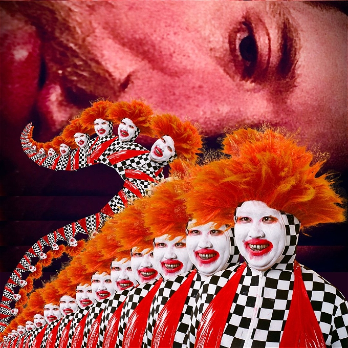 Su Mi is in white makeup in the foreground with a black and white checked jump suit and red latex scarf. Her hair is orange and her mouth red. A portion of Ciaran's face looms over her in the background.