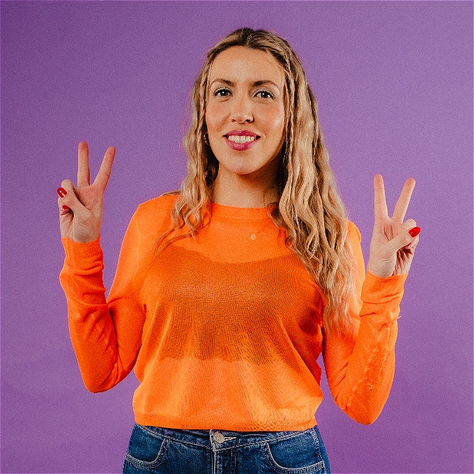 A smiling Lou Taylor holds both hands in a V shape with her fore and middle fingers in the peace sign