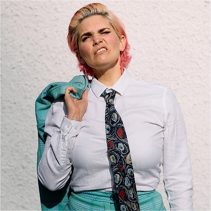 Female comedian wearing a suit, looking up and grimacing at the sun