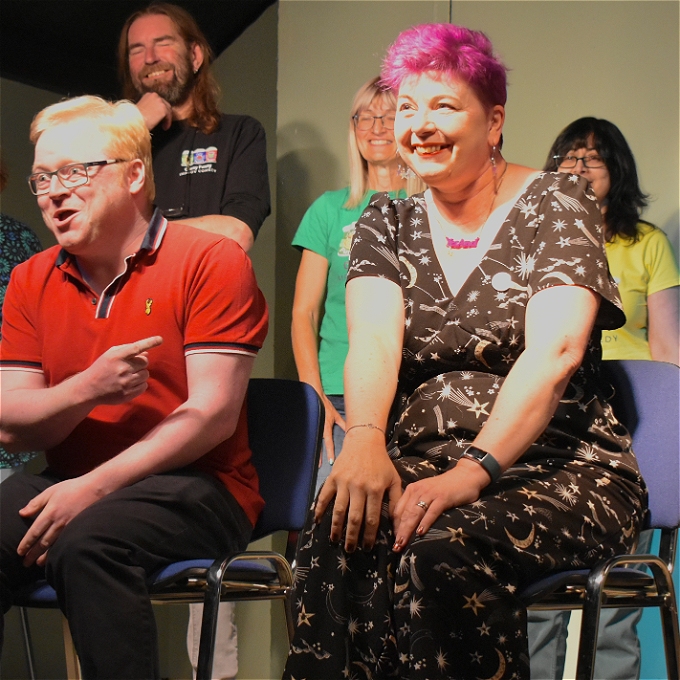 Female comedian with purple hair smiling and sitting next to blonde haired man pointing at her 3 other comedians in bright t shirts stood behind